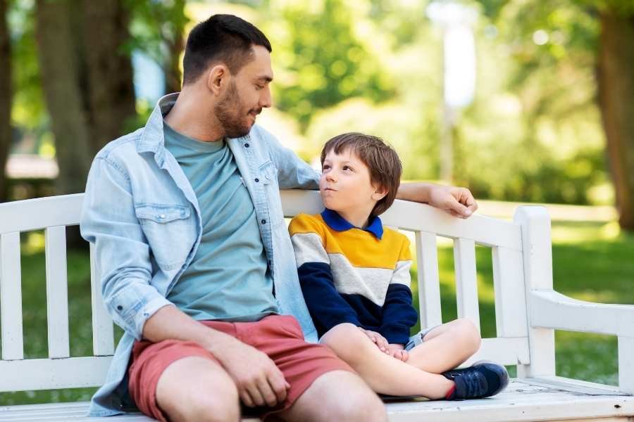Pai e um filho de cerca de 10 anos sentados em um banco conversando amigavelmente. Ilustração de como os pais podem ajudar a trabalhar a inteligência emocional.
